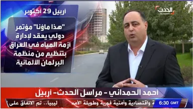 Television news frame of a man in a suit speaking outdoors, with Arabic text reading "أربيل 29 أكتوبر" and a headline about an international conference on Iraq's water crisis, plus a channel logo.