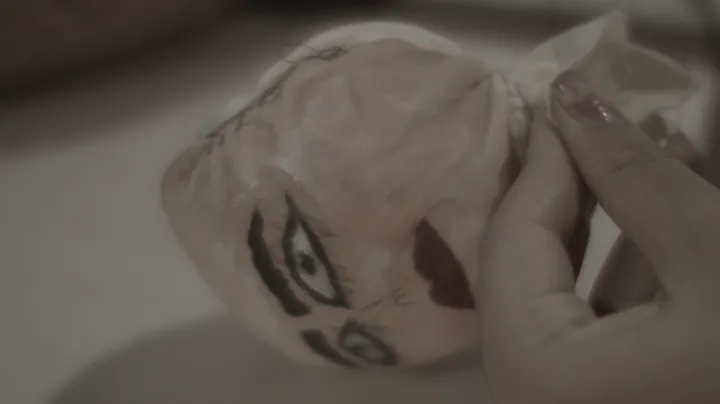 A hand with painted nails holds a fabric doll head with painted eyes, eyebrows and a red mark, against a plain background.