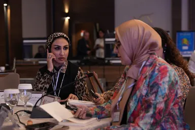 Two women sit at a table with water glasses and papers, one holding a phone to her ear while speaking, the other looking down at a phone.