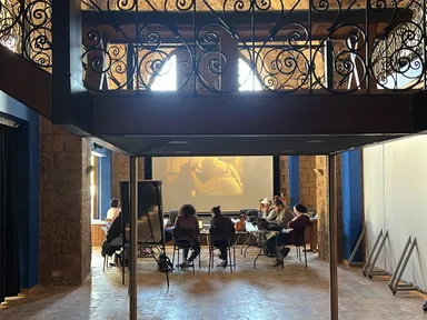 People seated around tables watching a projected video, viewed through an ornate wrought-iron balcony railing above.