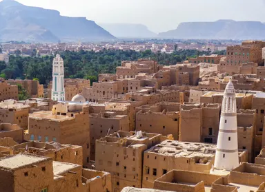 Dense cluster of sand-coloured mud-brick houses and towers with white minarets, backed by a green palm grove and distant mountains under a hazy sky.