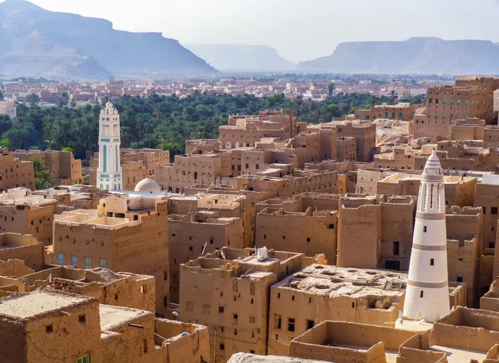 Dense cluster of sand-coloured mud-brick houses and towers with white minarets, backed by a green palm grove and distant mountains under a hazy sky.