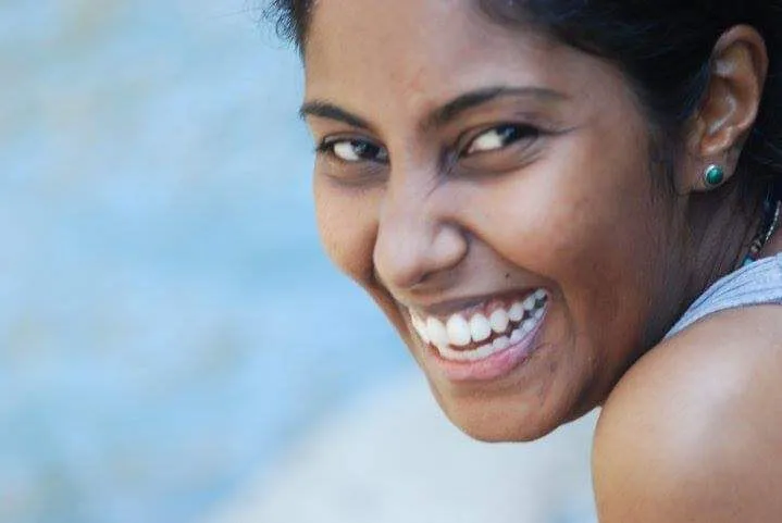 A woman looks back over her shoulder and smiles broadly outdoors, against a softly blurred light background.