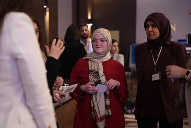 Three women stand together indoors, one in a red coat holding papers, another wearing a maroon jacket and a lanyard, with a screen and other people visible in the background.