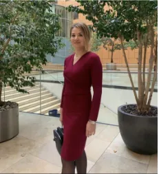 A woman in a maroon wrap dress stands smiling among potted trees in an indoor atrium, holding a bag.