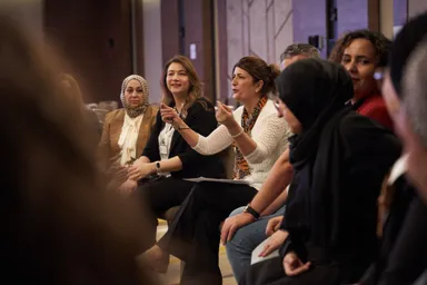 A group of women sit together in conversation, one gesturing while speaking, in an indoor venue.
