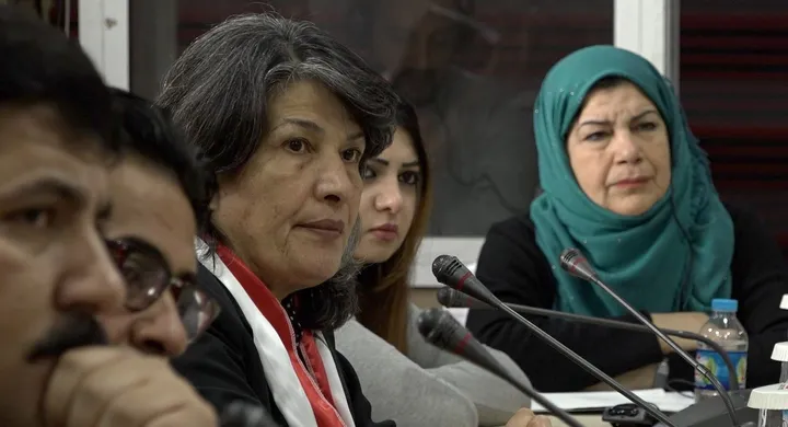 A group of people sits at a table with microphones, one woman wearing a teal headscarf.