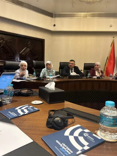 A group of people sit around a conference table in a meeting room, with a red flag and blue folders reading "elbarlament" on the table.