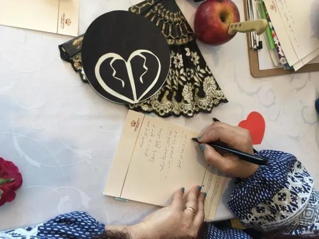 Overhead view of a hand writing Arabic text on a card beside a fan printed with a broken-heart symbol, an apple, a rose, and a red paper heart.