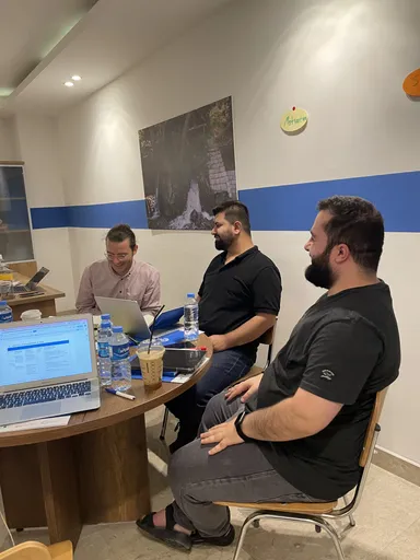 Three men sit at a round table with laptops and drinks in a room with a wall photo of a waterfall and a sticky note reading "Motivation".