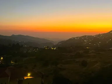 Hills and village lights beneath an orange sunset sky with a crescent moon.