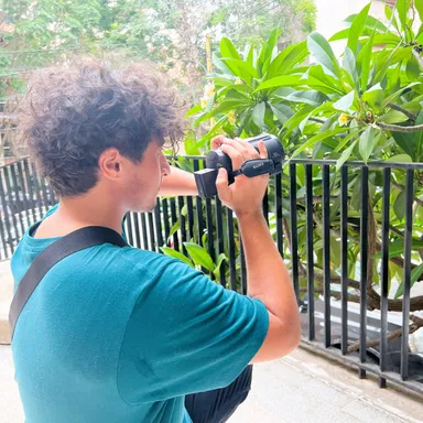 A person aims a video camera at green foliage behind a black metal fence.
