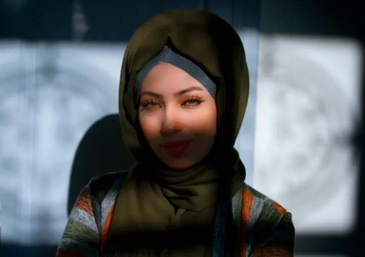 A woman in a dark green headscarf and a striped top smiles at the camera against a blurred patterned background.