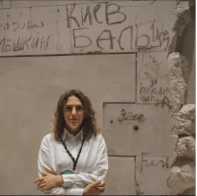 A woman with arms crossed stands in front of a wall inscribed with handwritten text, including "Киев" and "1945".