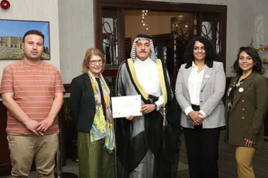 A group of five people pose together, one man holding a certificate, near a wall-mounted photo of ancient ruins and an open door.