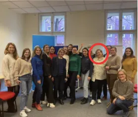 A group of about fifteen people pose together indoors for a photo, with one person's face circled in red.