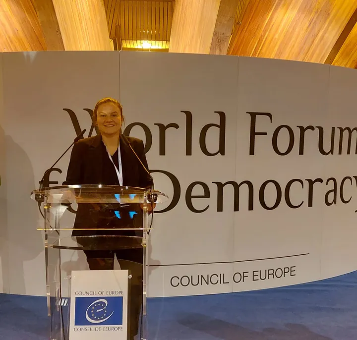 A woman standing at a podium in front of a wall reading "World Forum for Democracy" and "COUNCIL OF EUROPE".