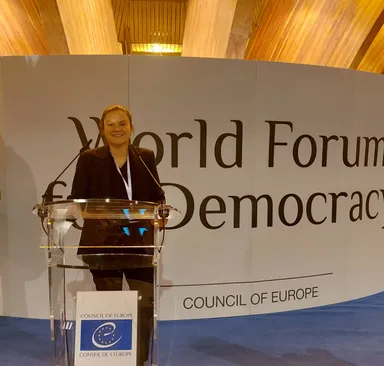 A woman standing at a podium in front of a wall reading "World Forum for Democracy" and "COUNCIL OF EUROPE".