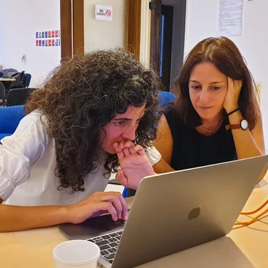 Two women lean over a laptop together indoors, one resting her chin on her hand.