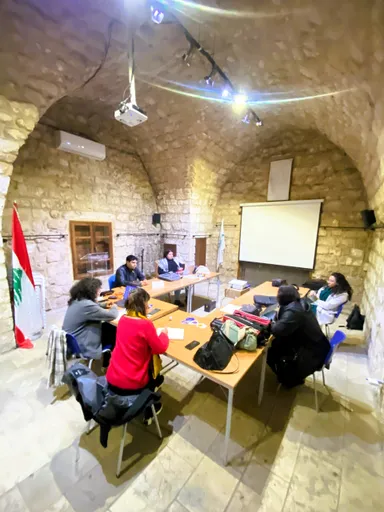 A group of people sit around tables in a vaulted stone room with a Lebanese flag and a blank projection screen.