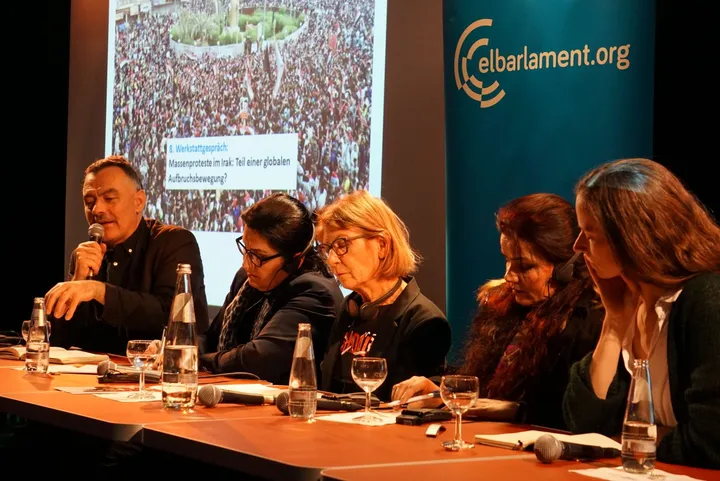Five people sit at a panel discussion table, a man speaking into a microphone, in front of a screen and a banner reading "elbarlament.org".