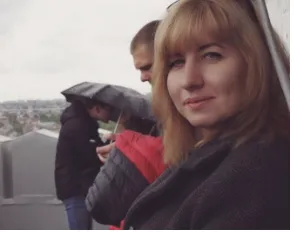 A woman looks toward the camera in the foreground while two people stand nearby under an umbrella on an outdoor rooftop under an overcast sky.