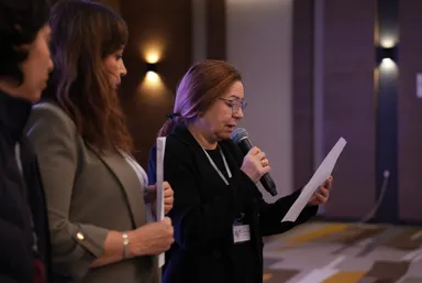 Three women stand together, one speaking into a microphone while reading from a paper she holds, with wall lights visible behind them.