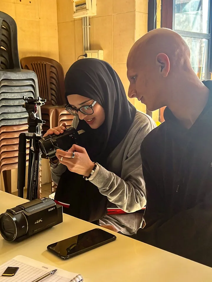 A woman wearing a hijab holds a video camera while a man beside her looks at its screen, with a second camcorder marked "SONY 4K" and a phone on the table.