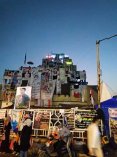 A structure covered in posters and string lights stands over a street market at dusk, with people passing stalls and a portrait display below.