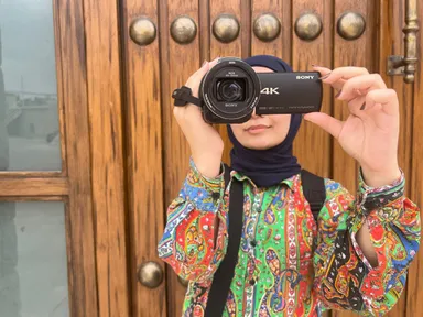 A woman in a headscarf looking through a Sony 4K camcorder held up to her eye, in front of an ornate wooden door.