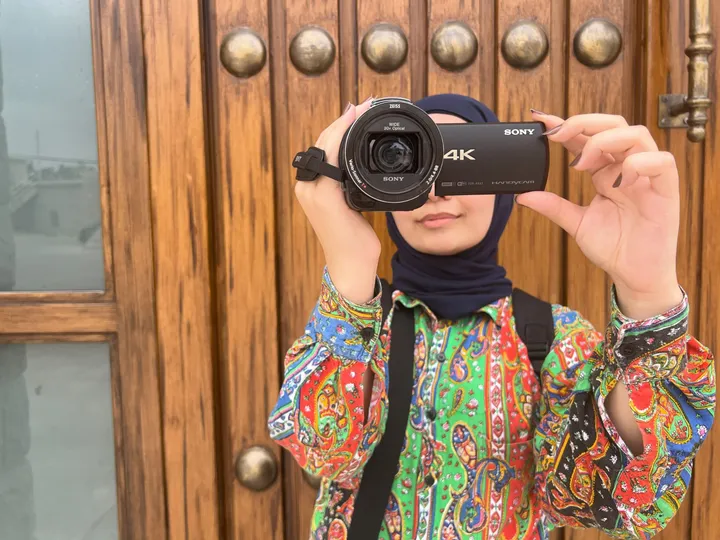 A woman in a headscarf looking through a Sony 4K camcorder held up to her eye, in front of an ornate wooden door.