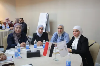 Four women in headscarves sit at a round table with water bottles, an Iraqi flag and a flag reading "CORAL".
