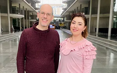 A man and a woman stand smiling in a glass-walled atrium lined with pillars and balconies.