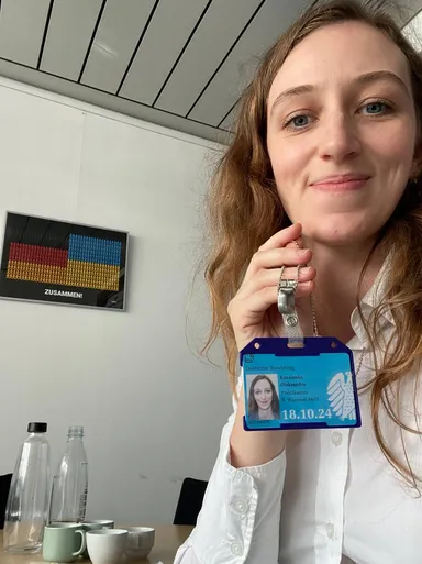 A woman holds up a "Deutscher Bundestag" visitor badge with a photo ID, in front of a poster reading "ZUSAMMEN!" in German and Ukrainian flag colors.