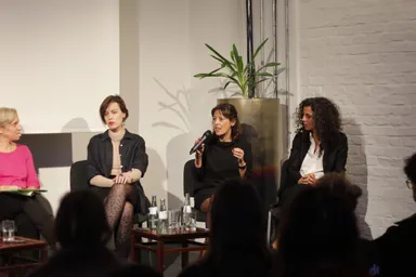 Four people sit on a panel stage in front of a potted plant and a white brick wall, a woman in the middle speaking into a microphone, water bottles on a table in front.