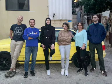 Six people standing in a row in front of a yellow car outside a building.
