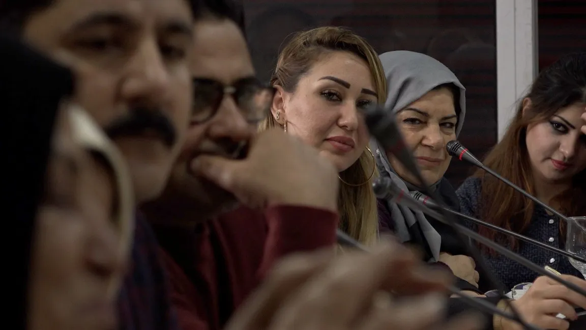 A group of people sit at a panel with microphones, including a woman in a grey headscarf.