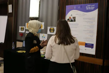 Two women wearing face masks talk in a conference hallway beside a poster reading "The War Made Me Stronger" in Arabic and English.