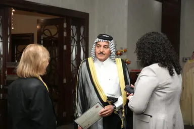 A man wearing a keffiyeh holds a certificate while speaking with two women, one holding a phone, near an open door.