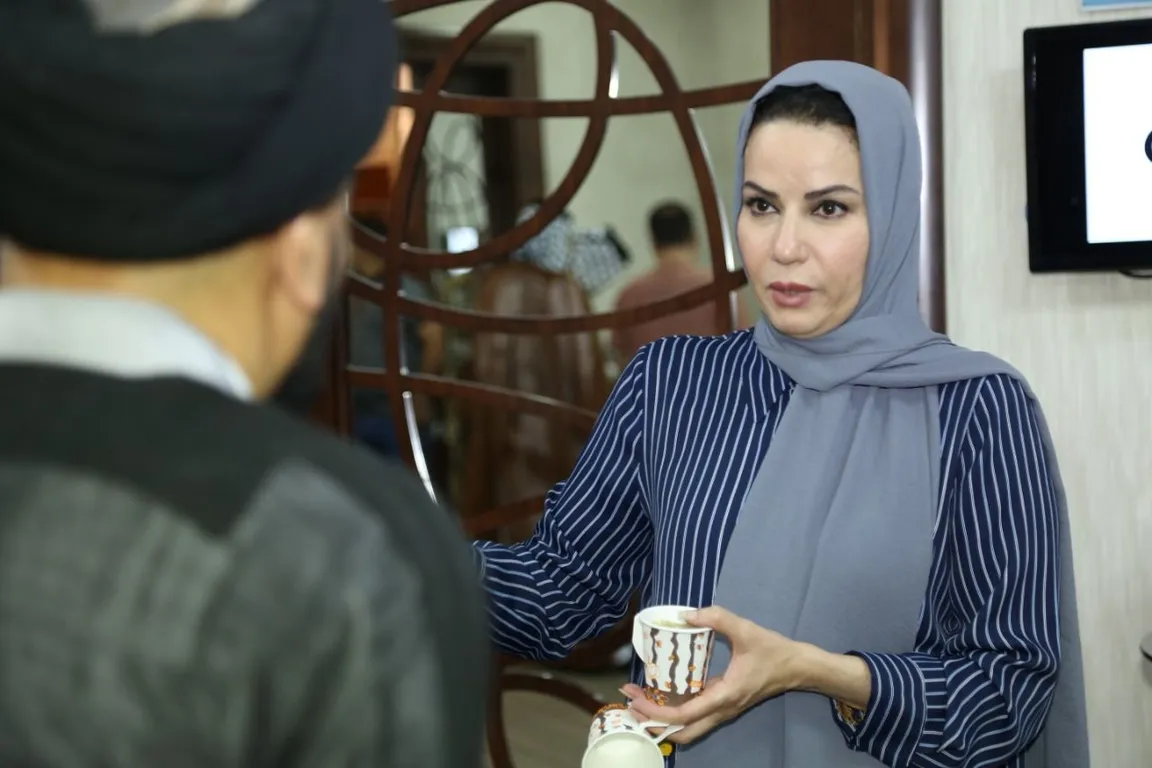 A woman wearing a headscarf holds a cup while speaking with a man in a black turban, seen from behind, near a wall-mounted screen.