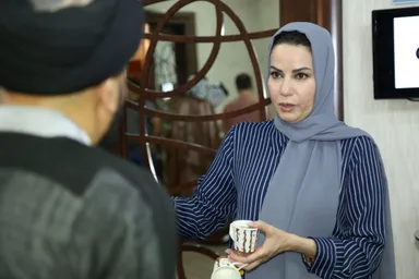 A woman wearing a headscarf holds a cup while speaking with a man in a black turban, seen from behind, near a wall-mounted screen.