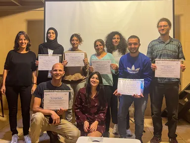 A group of people holding "Certificate of Participation" documents while posing together in front of a projection screen.