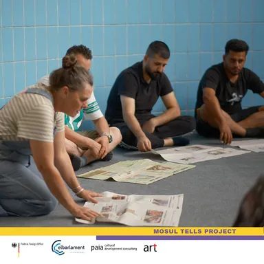 Four people sitting against a blue-tiled wall with newspaper pages spread on the floor in front of them, above a "MOSUL TELLS PROJECT" banner.