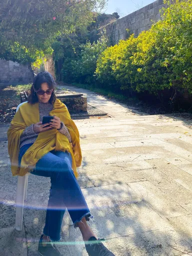 A woman in sunglasses and a yellow shawl sits on a chair outdoors looking at a phone, with greenery behind her.