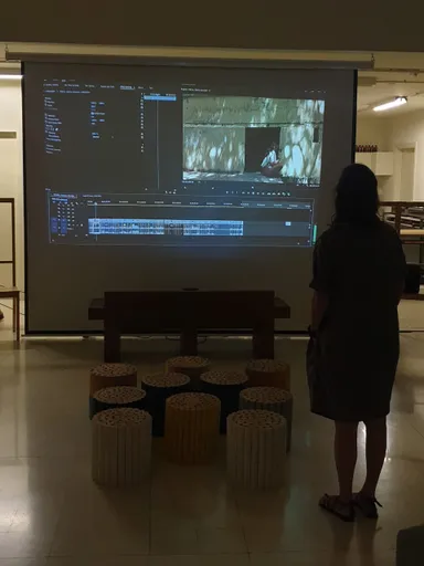 A person stands before a projection screen showing video-editing software, with round stools arranged in front.