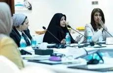 Three women sit at a conference table with microphones, water bottles, and papers in front of them.