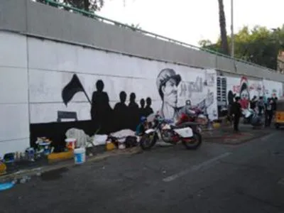 A wall mural shows silhouetted figures holding a flag beside a portrait of a man in a cap, with a motorcycle parked in front and people further along the street.