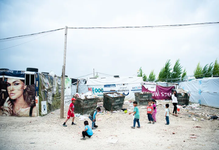 Children play on a dirt lot in an informal settlement of tents and containers, with a painted portrait of a woman's face on a structure in the background.