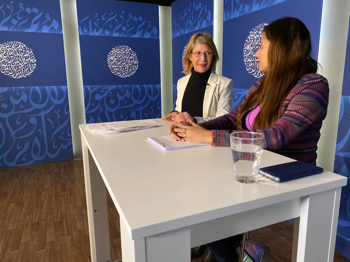 Two women sit talking at a white table in a TV studio with circular Arabic calligraphy panels behind them.