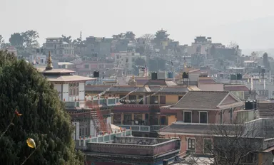 Aerial view of a dense hillside town with pagoda-roofed buildings and prayer flags, seen past a tree under a hazy sky.